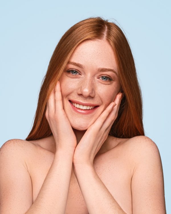 Beautiful young woman with hands on freckled cheeks smiling and looking at camera against blue background